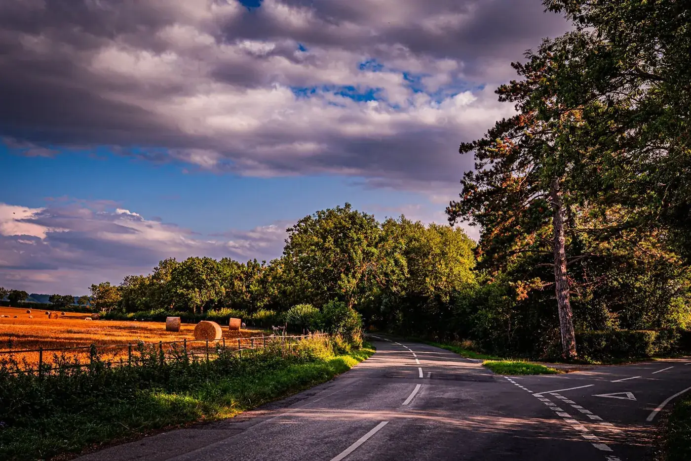 Country road winding past hay bales and trees under dramatic evening cloud