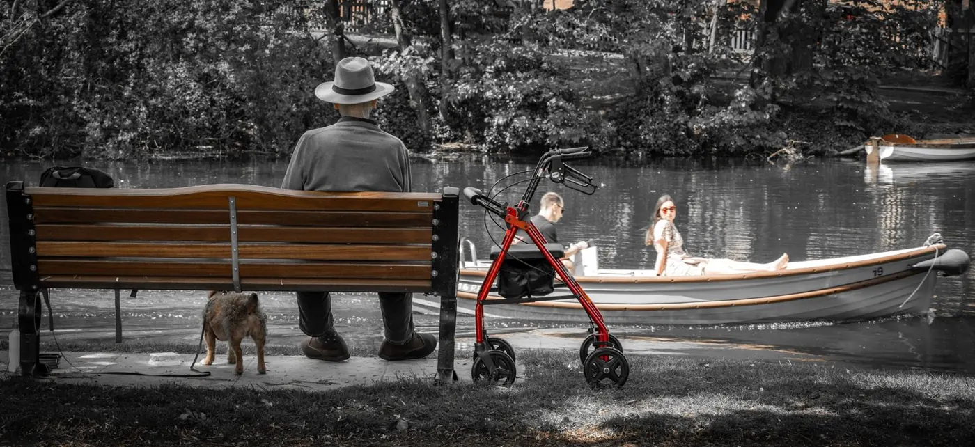An older man in a hat sits on a riverside bench with his dog and walking frame as a couple pass by boat