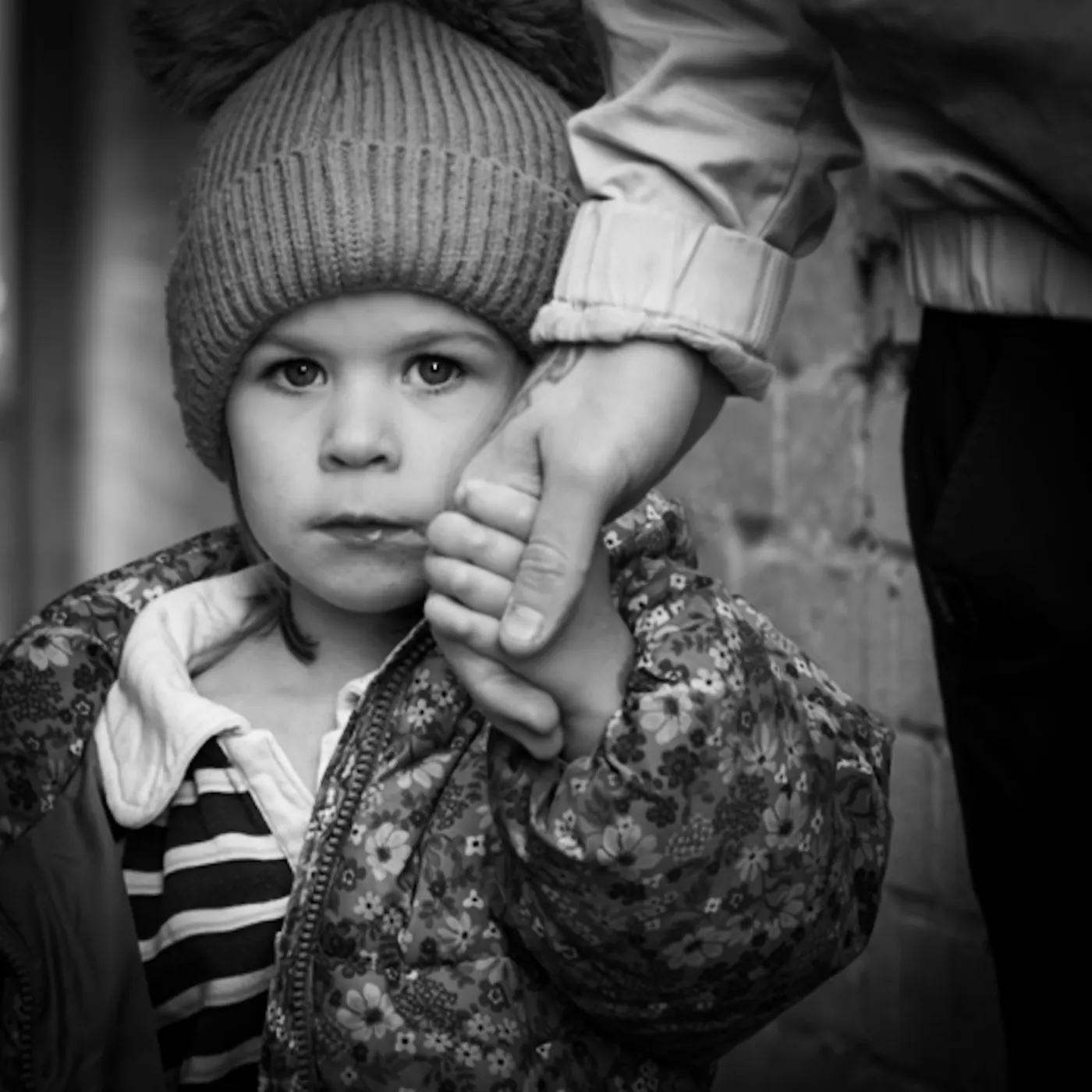Black and white street portrait of a young child in a bobble hat holding an adult's hand