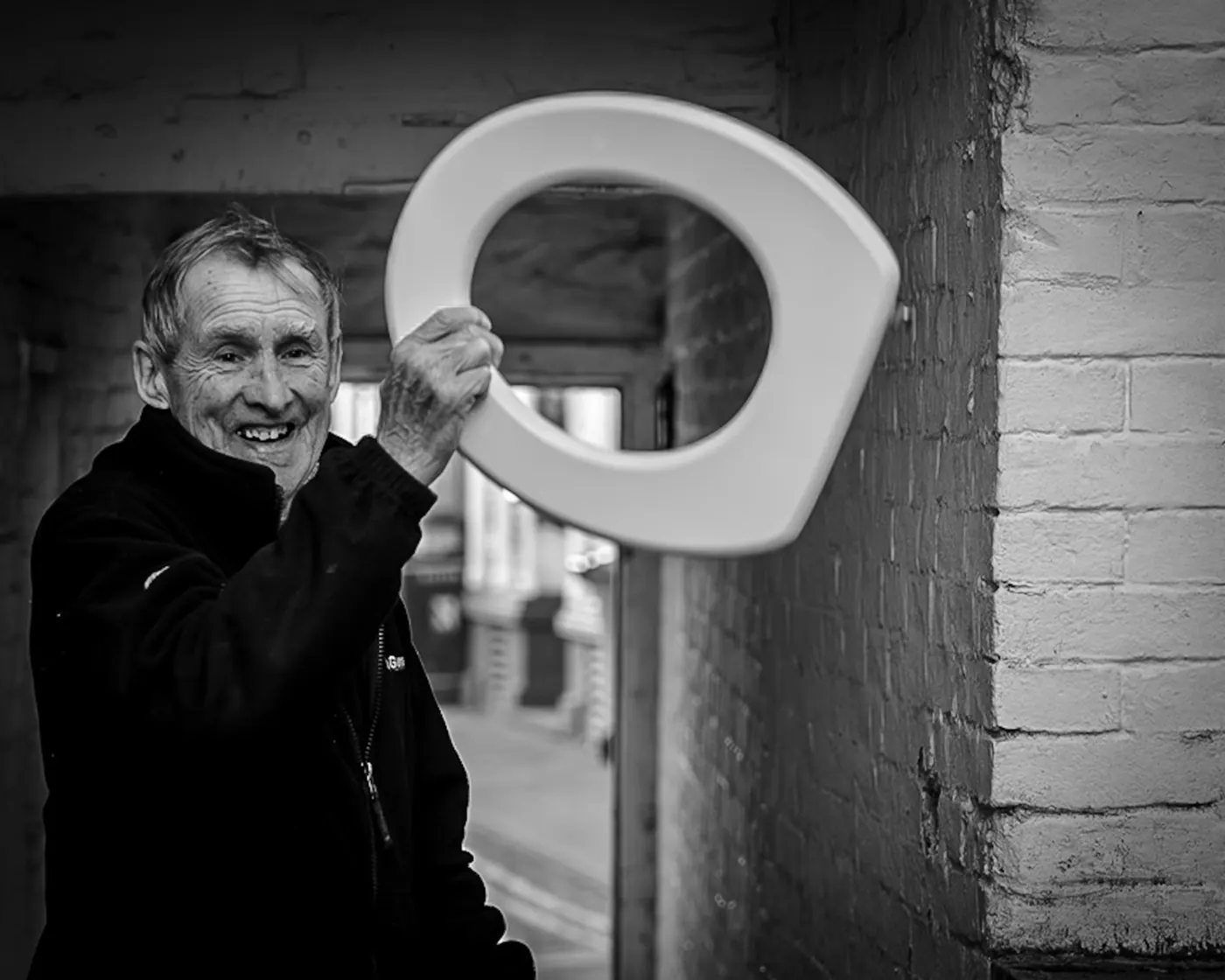 Black and white street portrait of a grinning older man holding up a white toilet seat in an alleyway