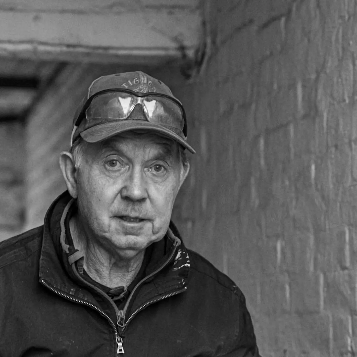 Black and white street portrait of an older man in a cap with safety glasses pushed up