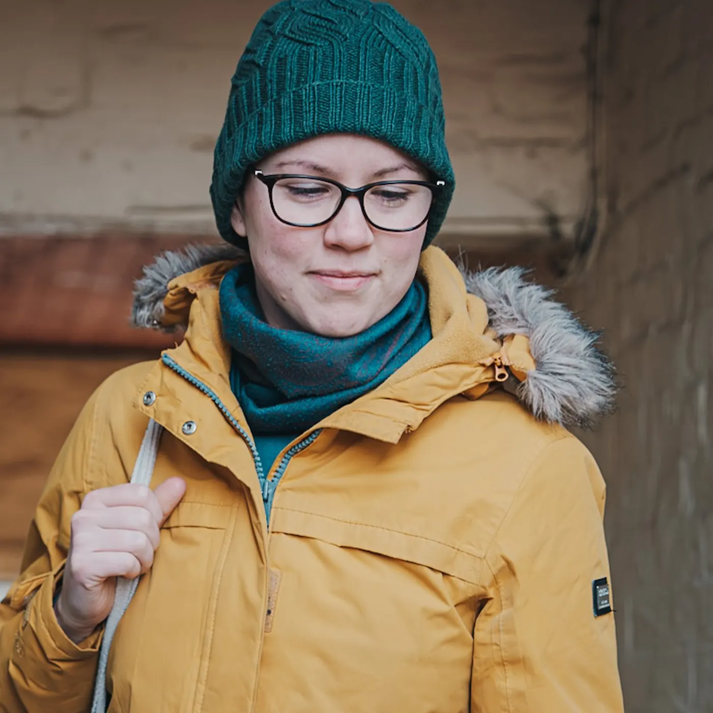 Street portrait of a woman in a green knitted hat, glasses and mustard coat, glancing down and smiling